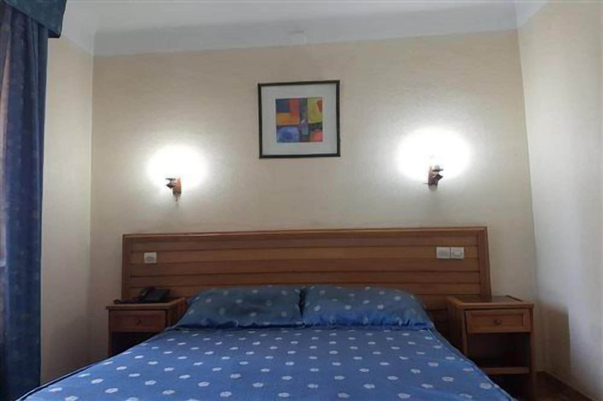 A bedroom with a wooden headboard, two bedside tables, a bed with a patterned bedspread, and two wall-mounted lights.