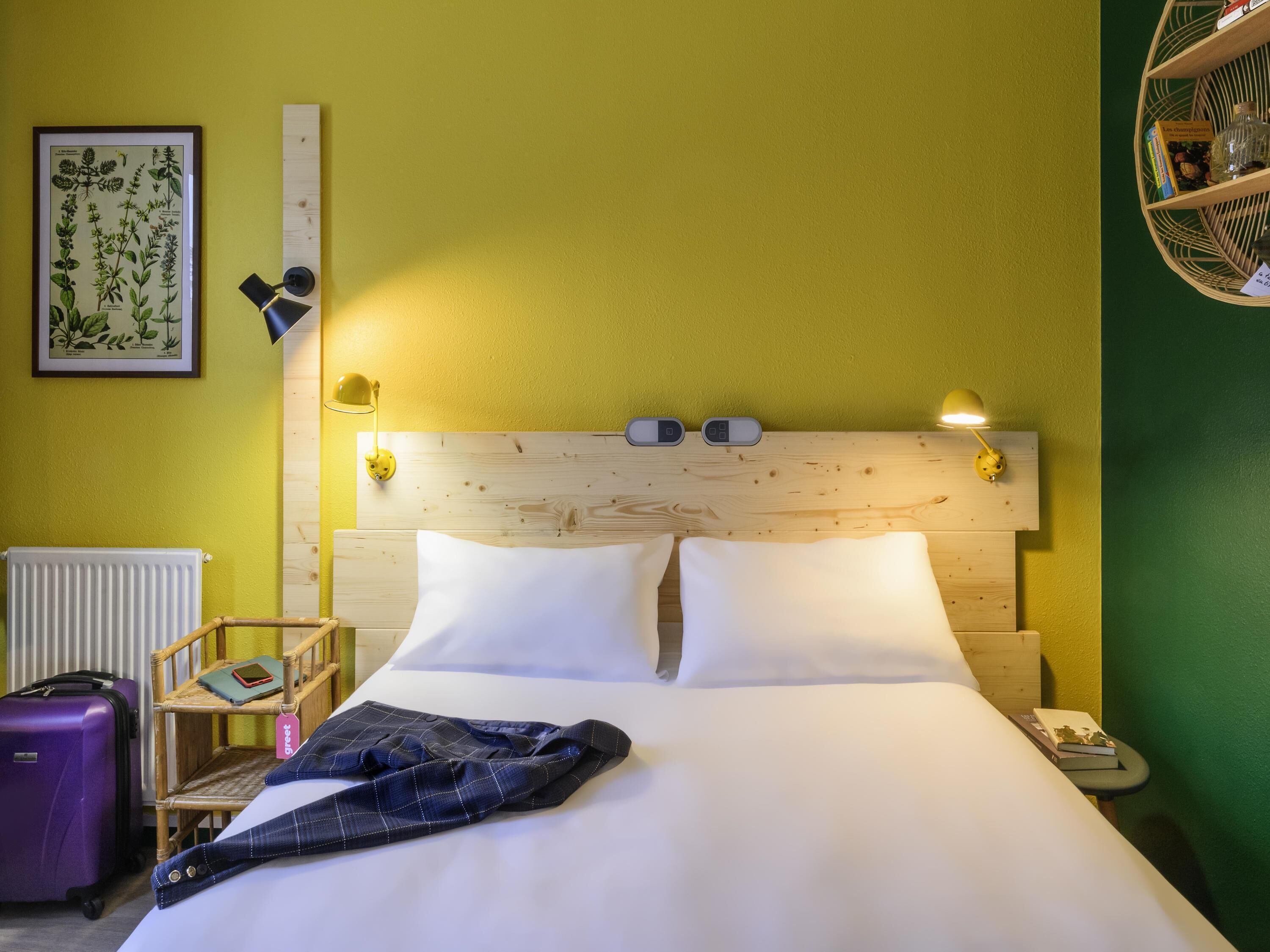 Una habitación de hotel con cabecera de madera, dos lámparas de noche, una ilustración botánica enmarcada, una mesita de noche con un libro y una botella, y una maleta morada.