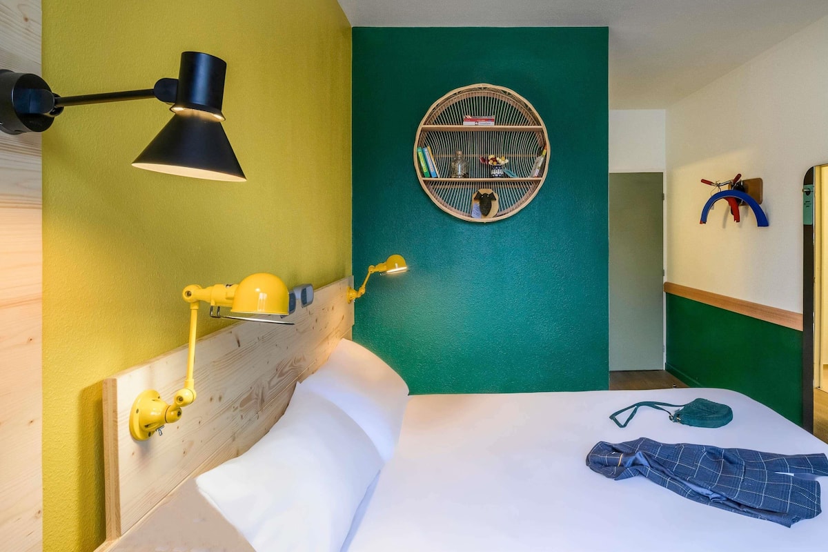 A room with a bed, a headboard, a wall-mounted lamp, a circular shelf with books and decorative items, and a door with a towel rack.