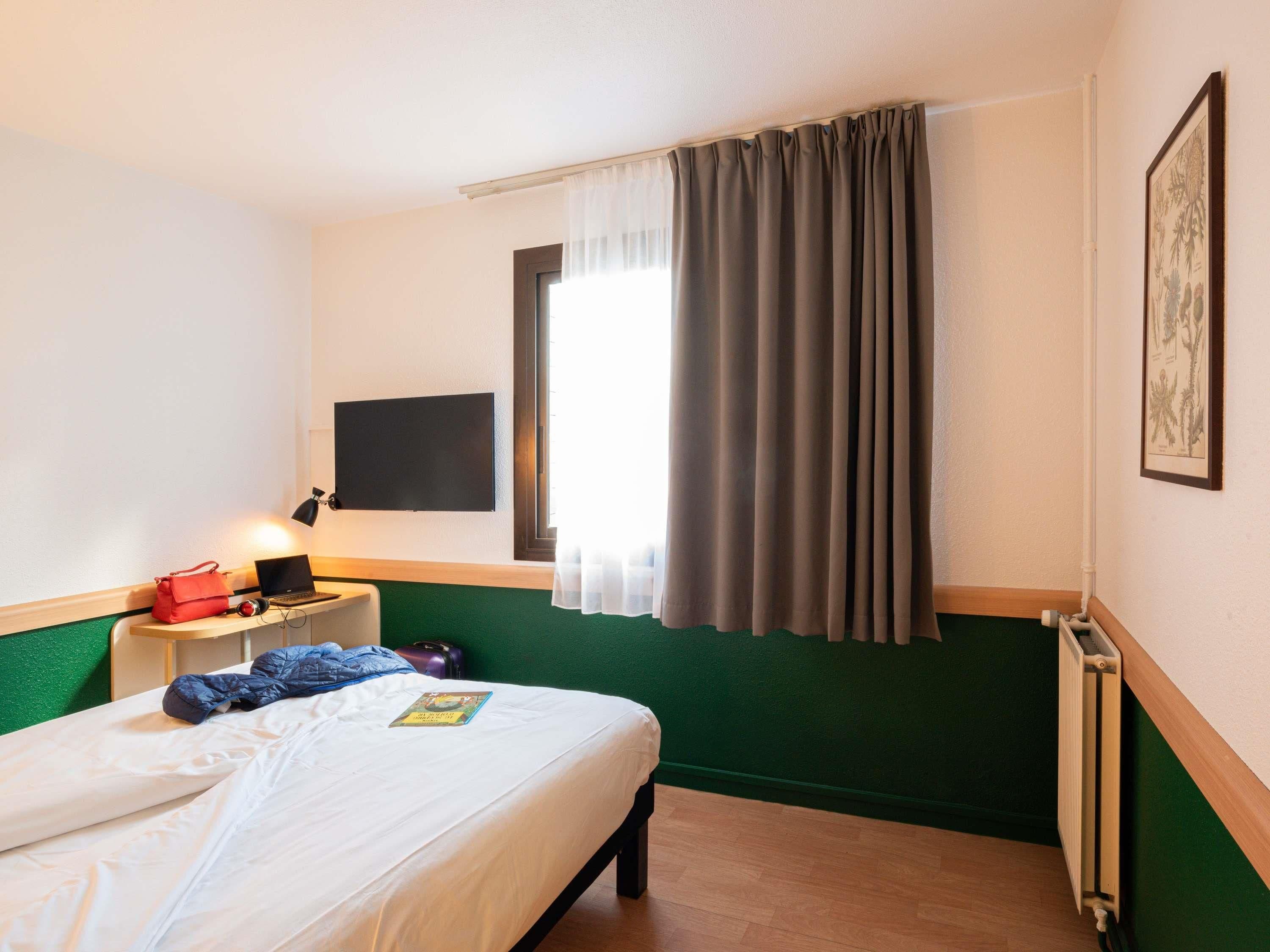 Een hotelkamer met een bed, een bureau met een laptop, een rode tas en een lijst in een lijstwerk.