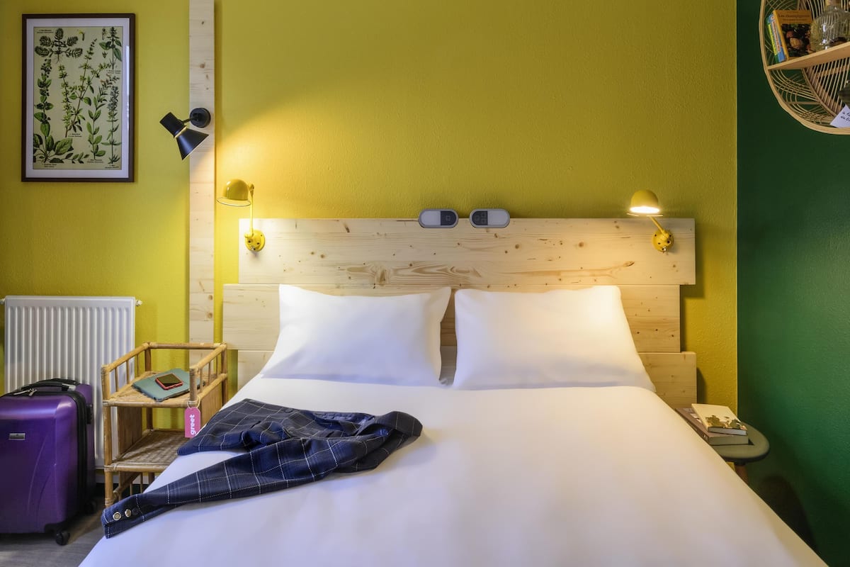 Hotellihuone, jossa on puinen sängynpääty, kaksi yöpöytävalaisinta, kehystetty kasvitieteellinen kuva, pieni yöpöytä, jossa on kirja ja pullo, sekä violetti matkalaukku.