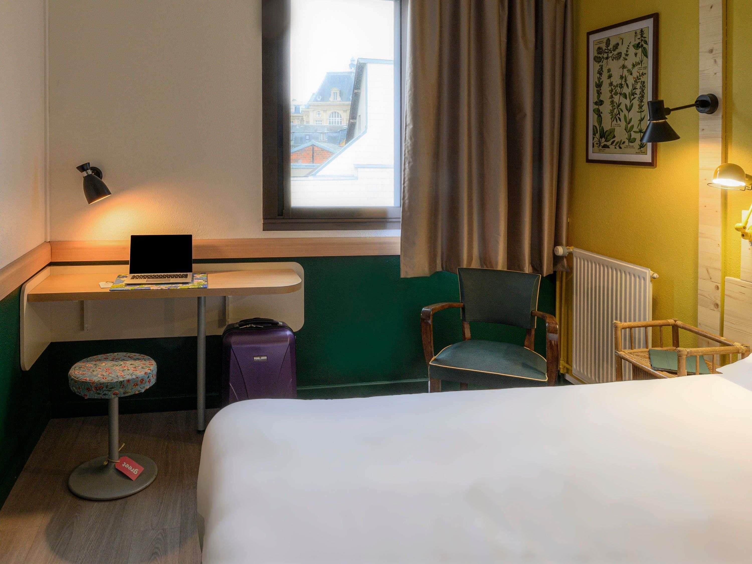 A hotel room with a bed, a desk with a laptop, a chair, a suitcase, and a window with curtains.