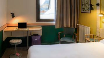 A hotel room with a bed, a desk with a laptop, a chair, a suitcase, and a window with curtains.