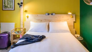 A hotel room with a wooden headboard, white bedding, a small bedside table with a lamp, a framed botanical print on the wall, and a suitcase on the floor. - Greet Hotel Evreux Centre - Route de Normandie by Accor (Evreux)