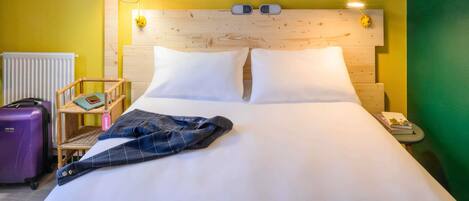 Une chambre d’hôtel avec une tête de lit en bois, du linge de lit blanc, une petite table de chevet avec une lampe, une estampe botanique encadrée au mur et une valise posée sur le sol.