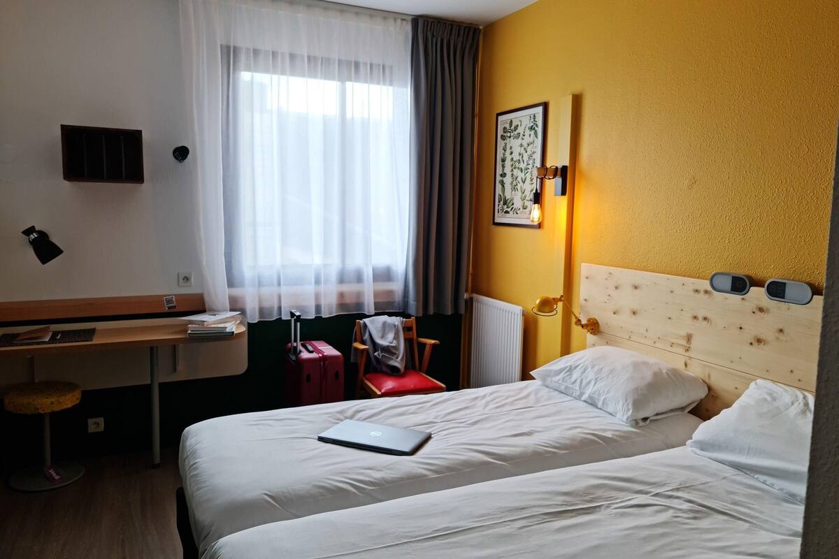 A hotel room with a bed, a desk, a chair, a suitcase, a lamp, and a wall-mounted light fixture.