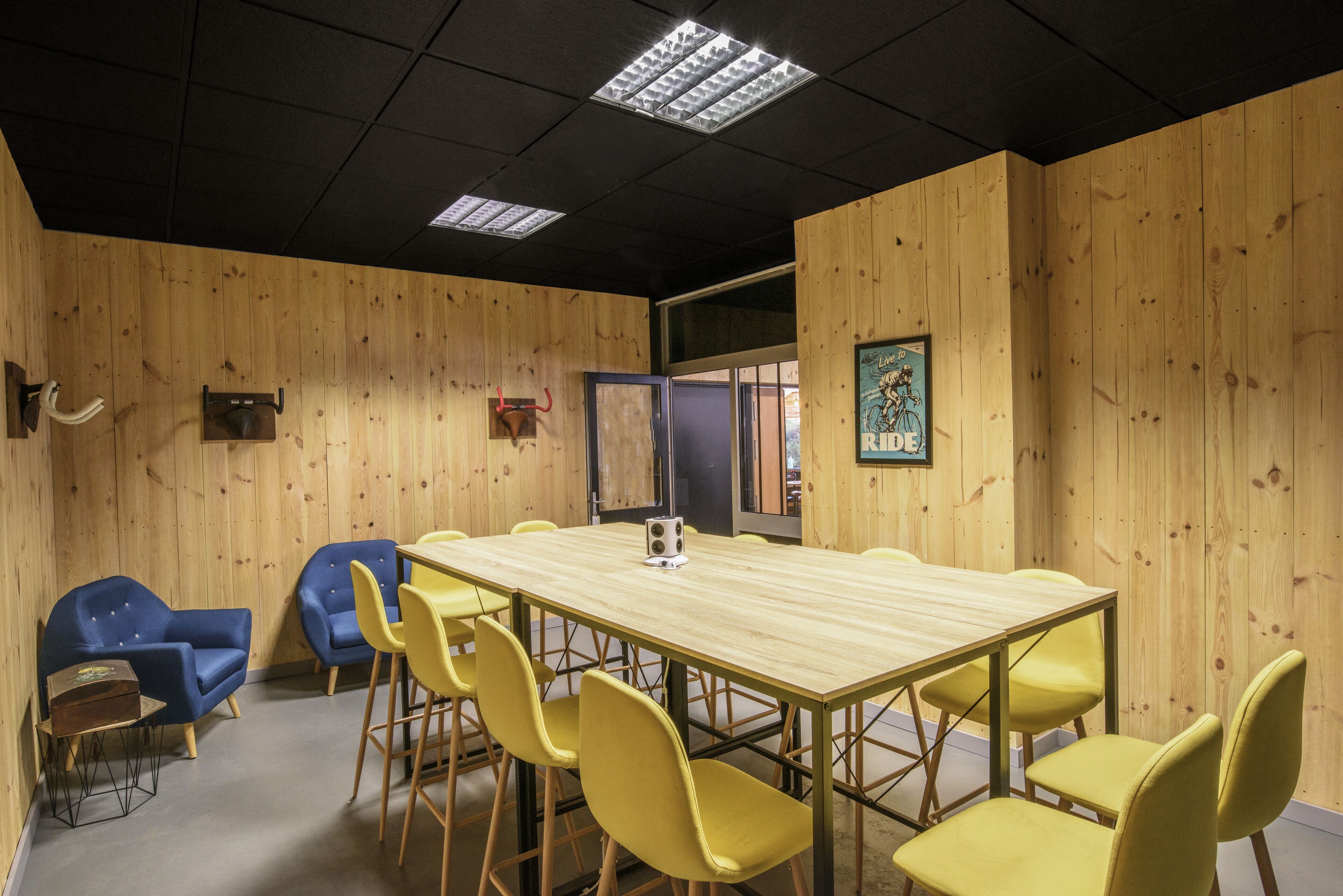 Un salón de conferencias con una mesa de madera y sillas amarillas, un sofá azul y un techo negro con luces fluorescentes.