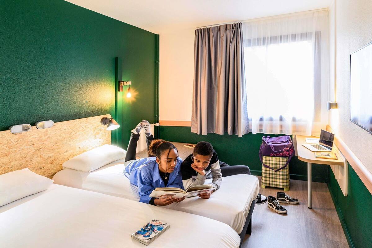 A hotel room with two beds, a desk with a laptop, a window with curtains, and a green wall.