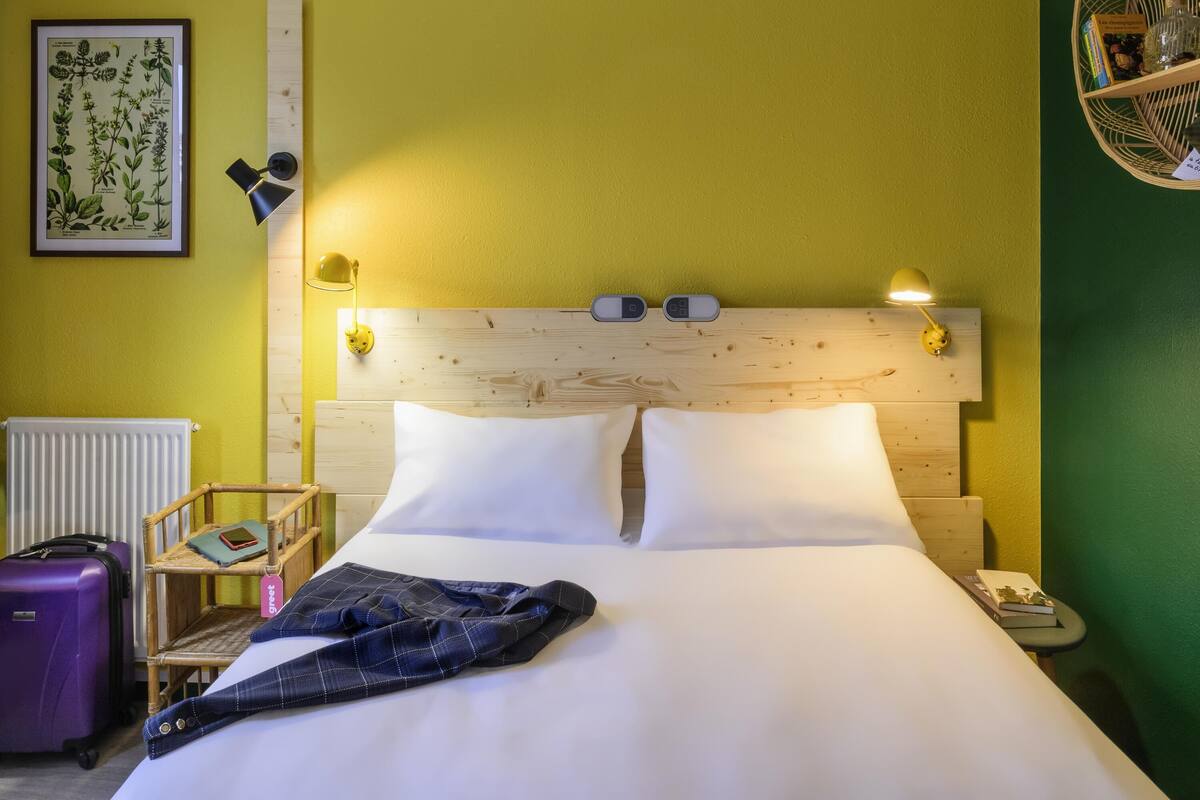 A hotel room with a wooden headboard, two bedside lamps, a framed botanical illustration, a small bedside table with a book and a bottle, and a purple suitcase.