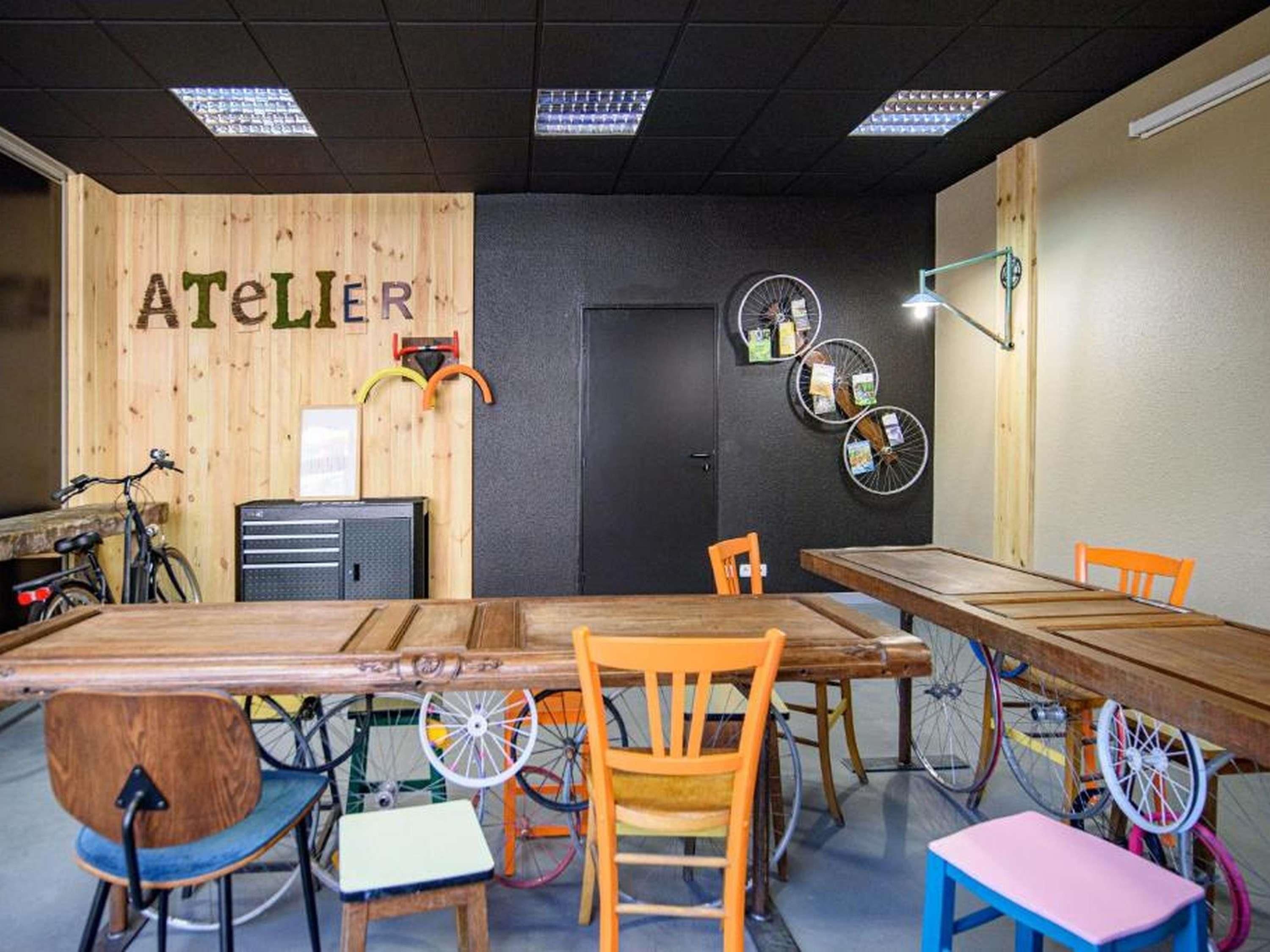 A room with a long wooden table, multiple chairs, bicycles, and a wall with the word 'ATELIER'.
