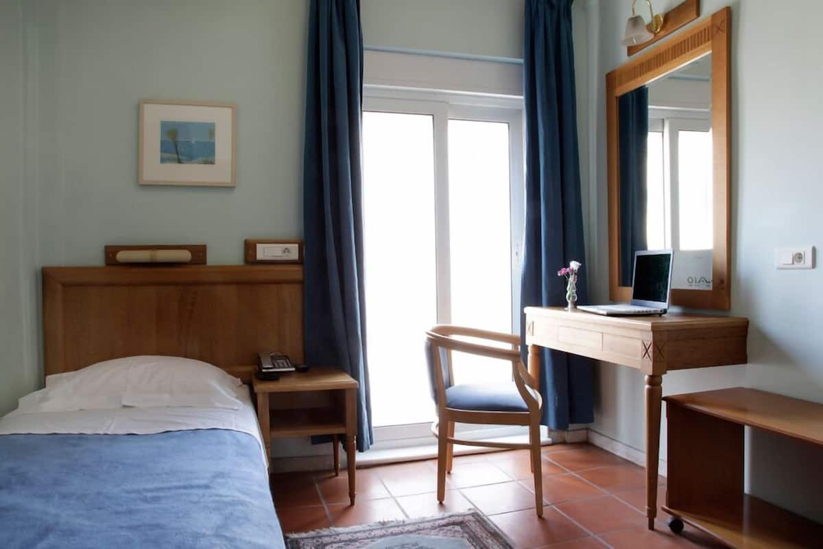 A bedroom with a bed, a desk with a laptop, a chair, a small table, a mirror, and a picture on the wall.