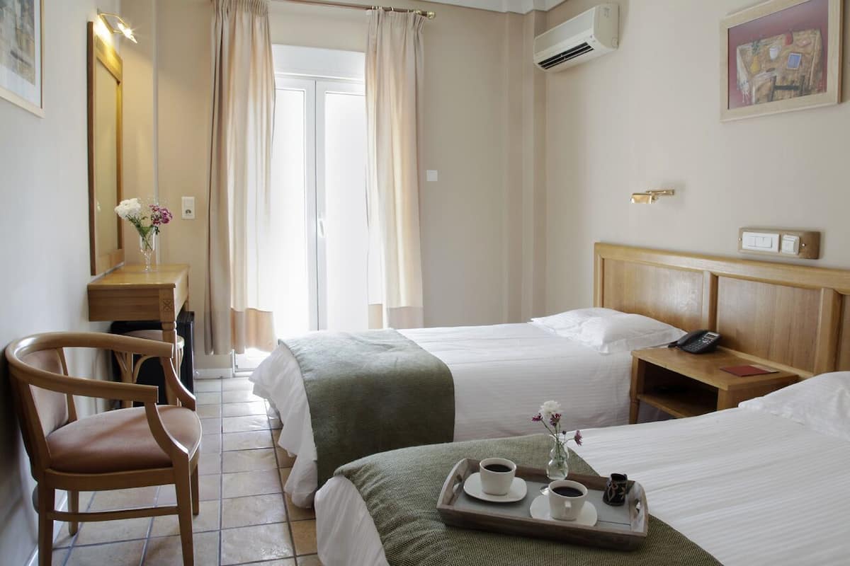 A hotel room with two beds, a wooden chair, a desk with a vase of flowers, and a tray with cups and a teapot.
