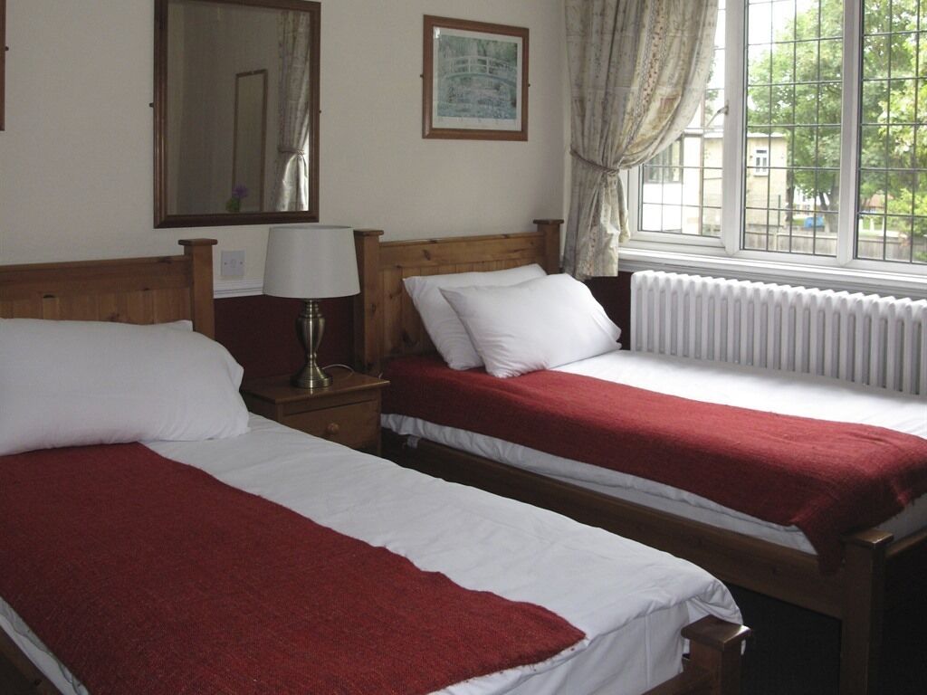 Two single beds with red blankets, a wooden headboard, a nightstand with a lamp, a window with a view of buildings, and a radiator.