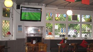 Un restaurante con muebles de madera, un televisor mostrando un partido deportivo y banderas colgadas del techo.