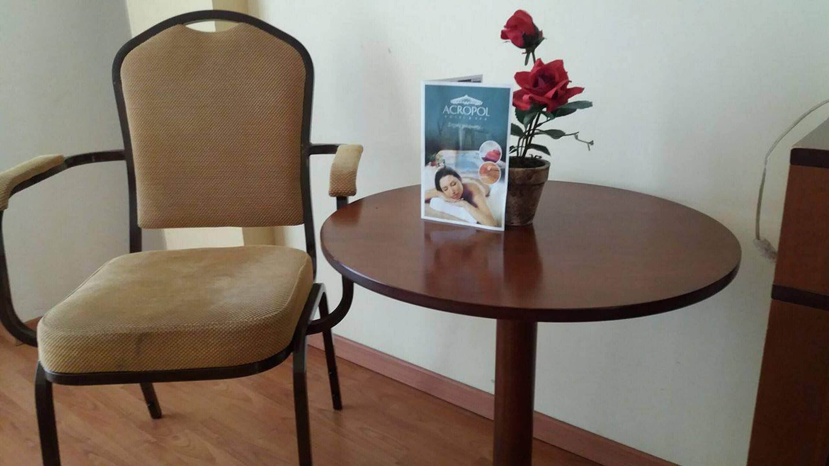 Una silla con respaldo tejido y asiento acolchado, una mesa redonda de madera con un folleto y una planta en maceta con flores rojas, y un piso de madera.