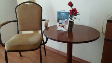 Una silla con respaldo tejido y asiento acolchado, una mesa redonda de madera con un folleto y una planta en maceta con flores rojas, y un piso de madera.