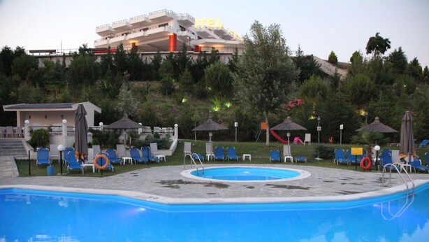 A swimming pool with lounge chairs and umbrellas, a hotel building in the background, and a hilly landscape.