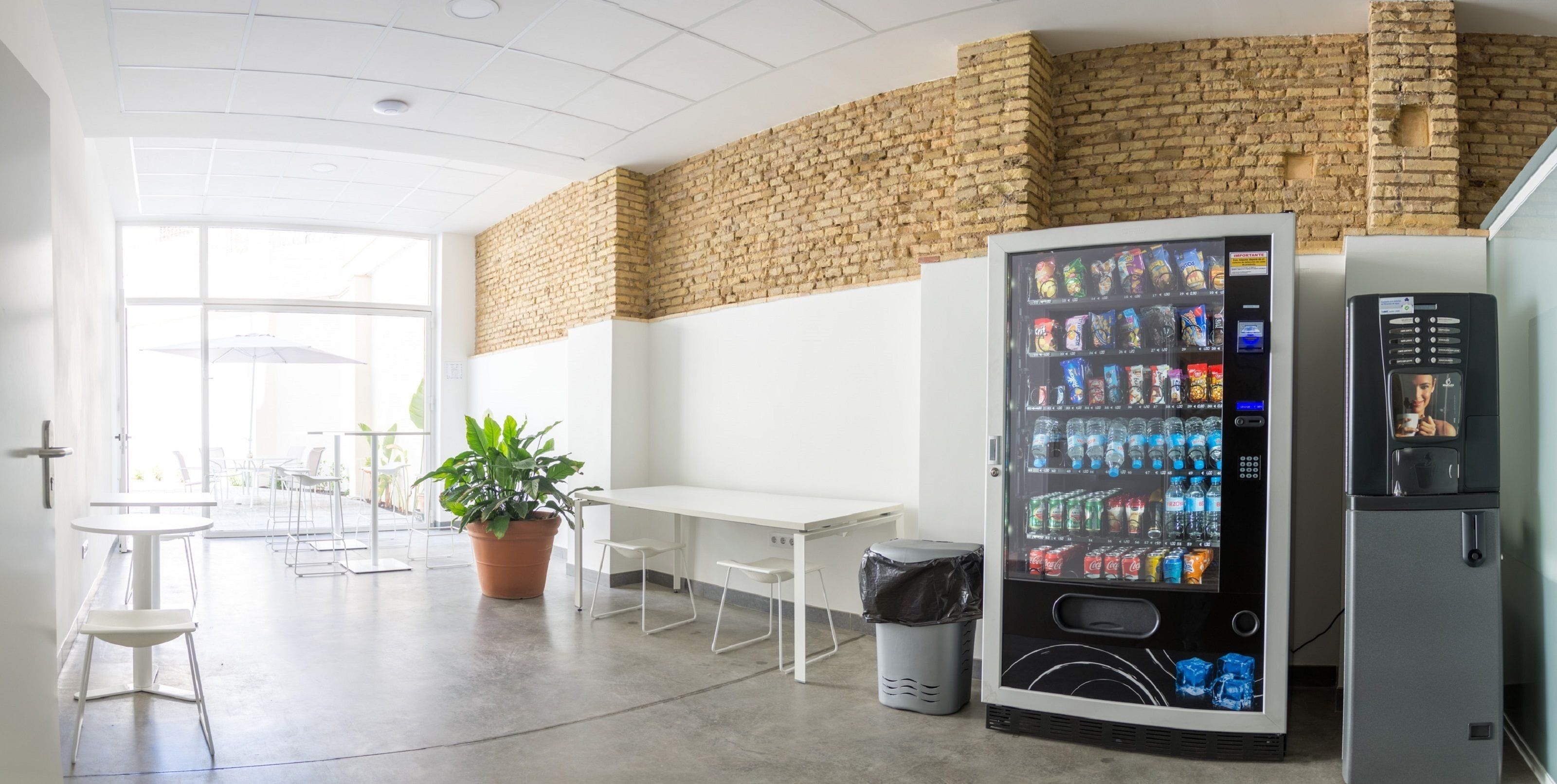 A modern indoor space with a brick wall, a vending machine, a trash bin, and a potted plant.