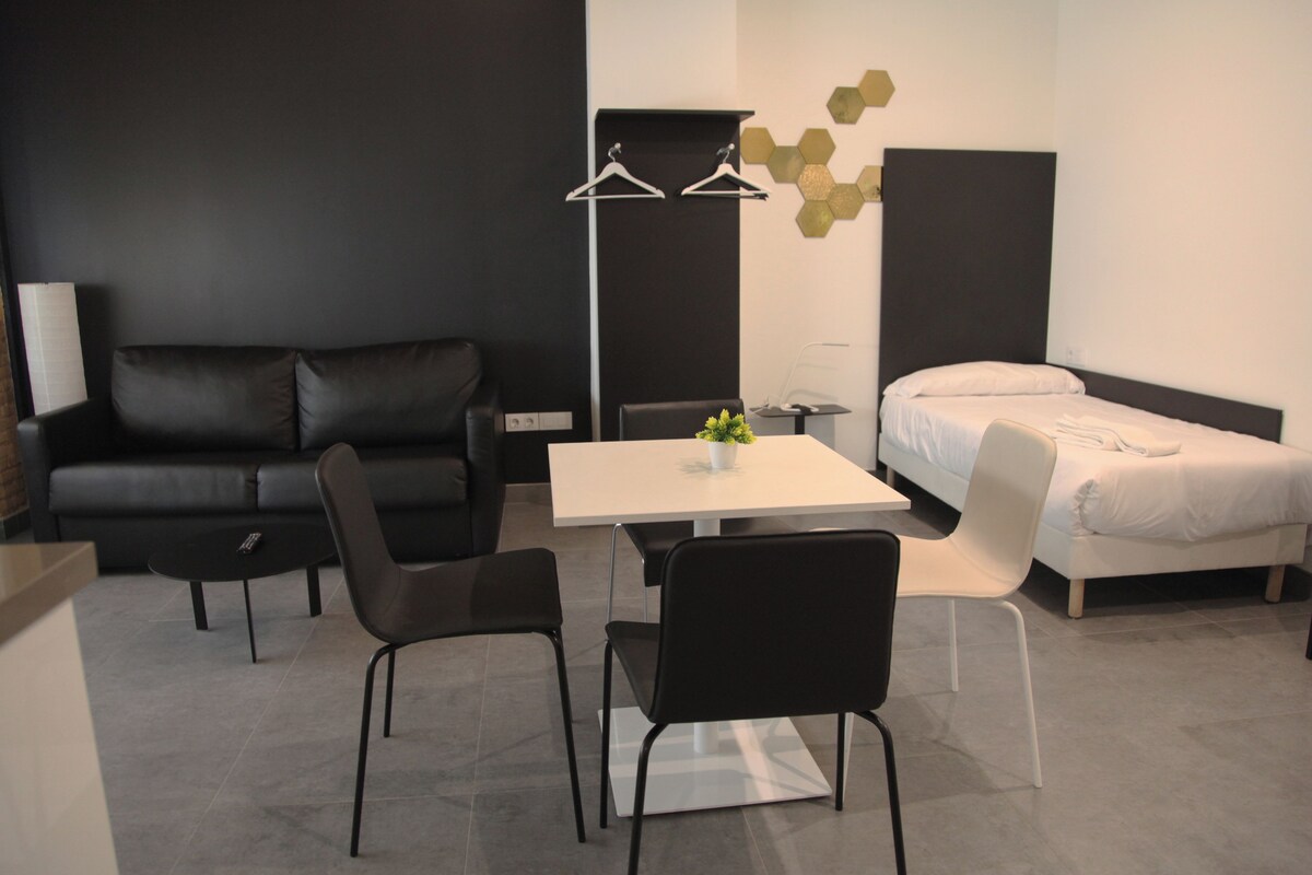 A modern hotel room with a black leather sofa, a white bed, a small table with chairs, and a bedside table with a lamp.