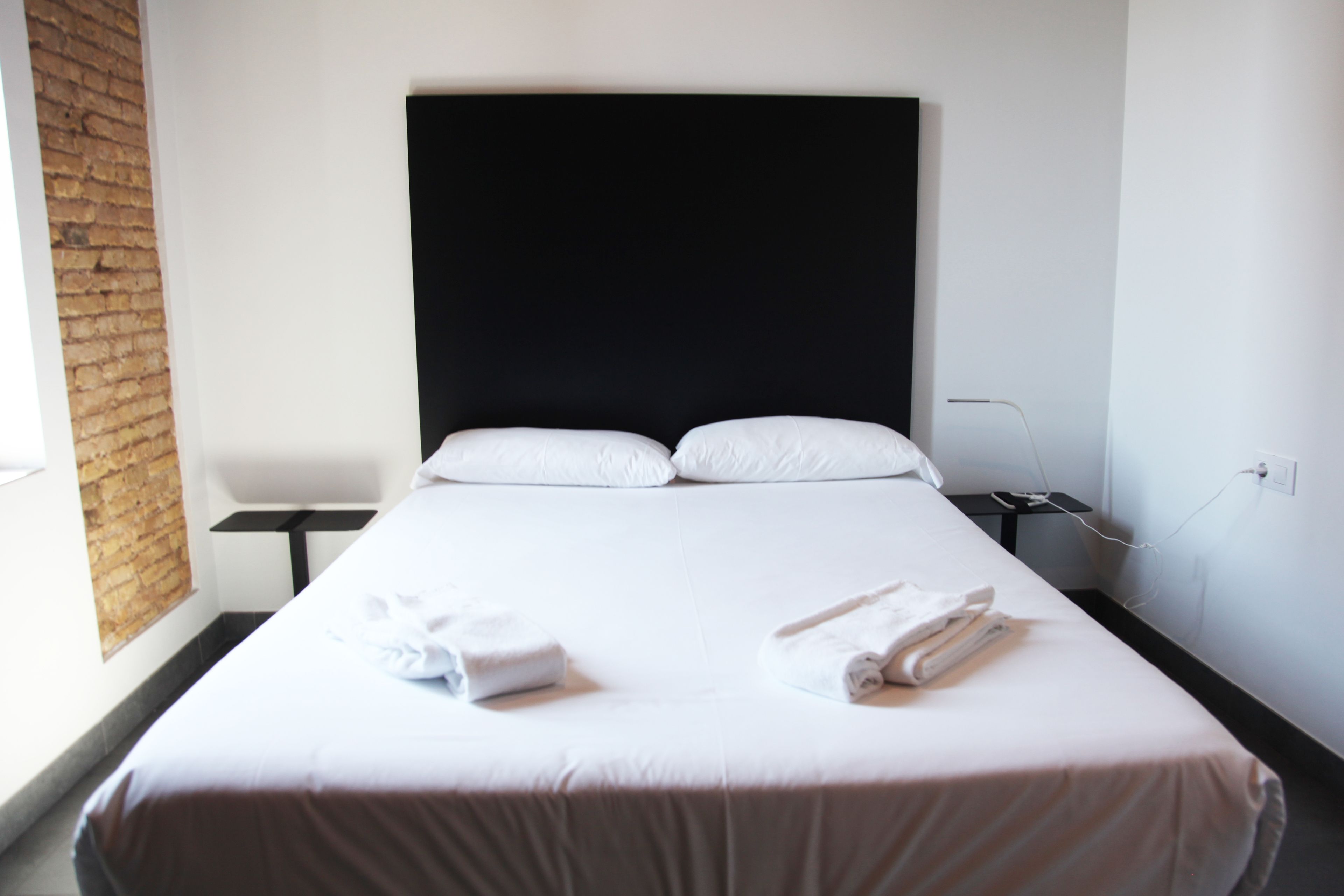 Una habitación de hotel con una cama grande, cabecera negra, ropa de cama blanca y una pared de ladrillo.