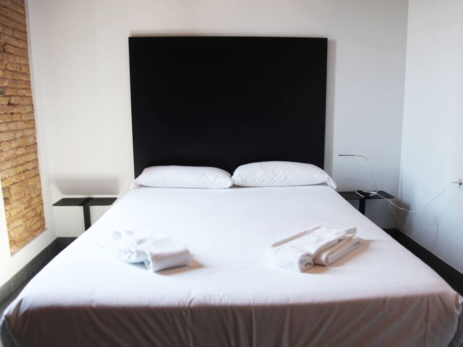Una habitación de hotel con una cama grande, cabecera negra, ropa de cama blanca y una pared de ladrillo.