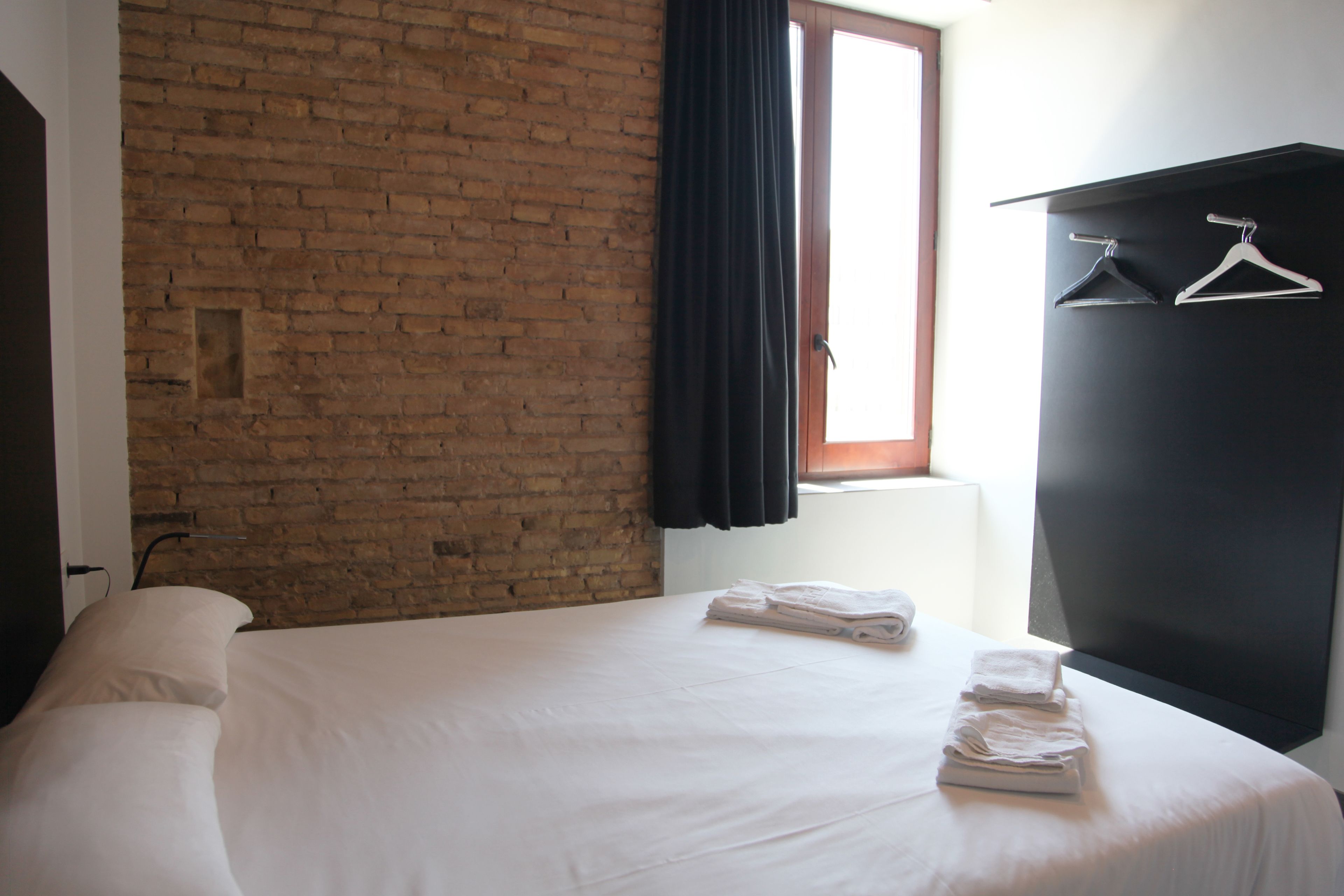 Een hotelkamer met een bakstenen muur, een bed met witte lakens, een raam met zwarte gordijnen en een zwarte garderobe met kleerhangers.
