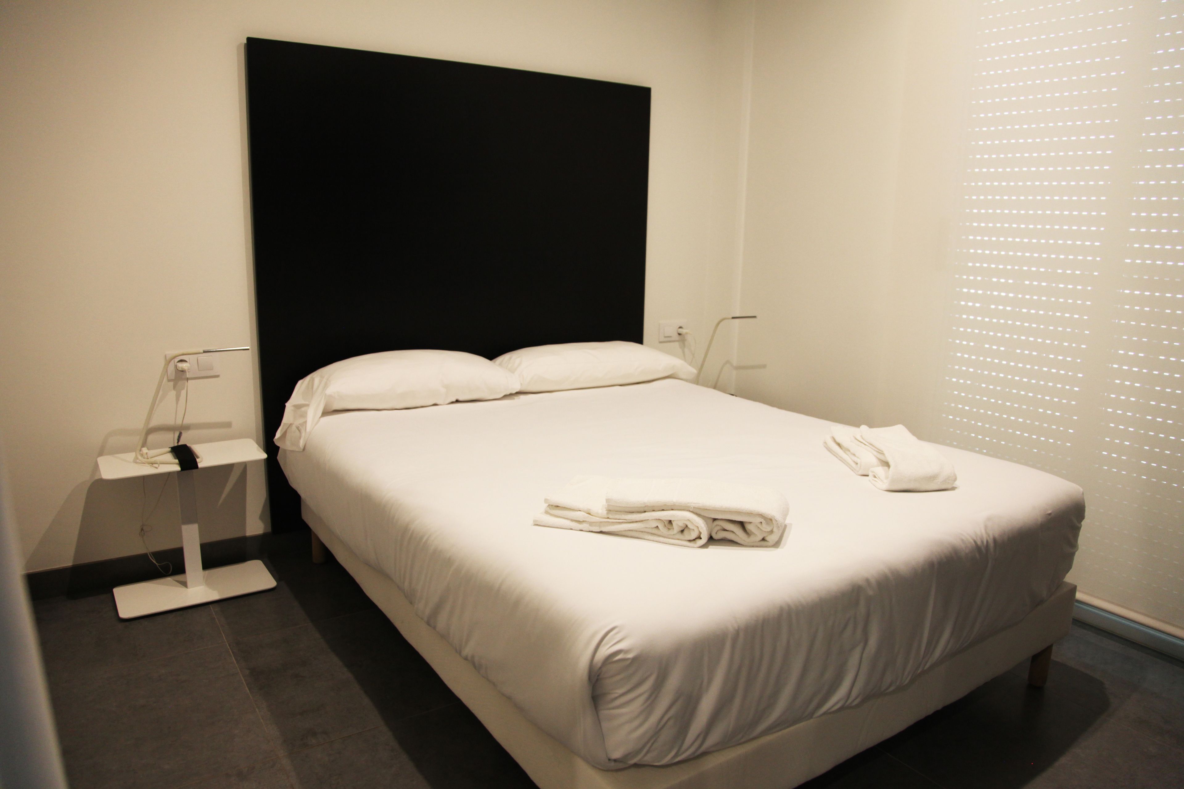 Una cama bien hecha con ropa de cama y toallas blancas, una mesita de noche con lámpara, y una ventana con persianas.