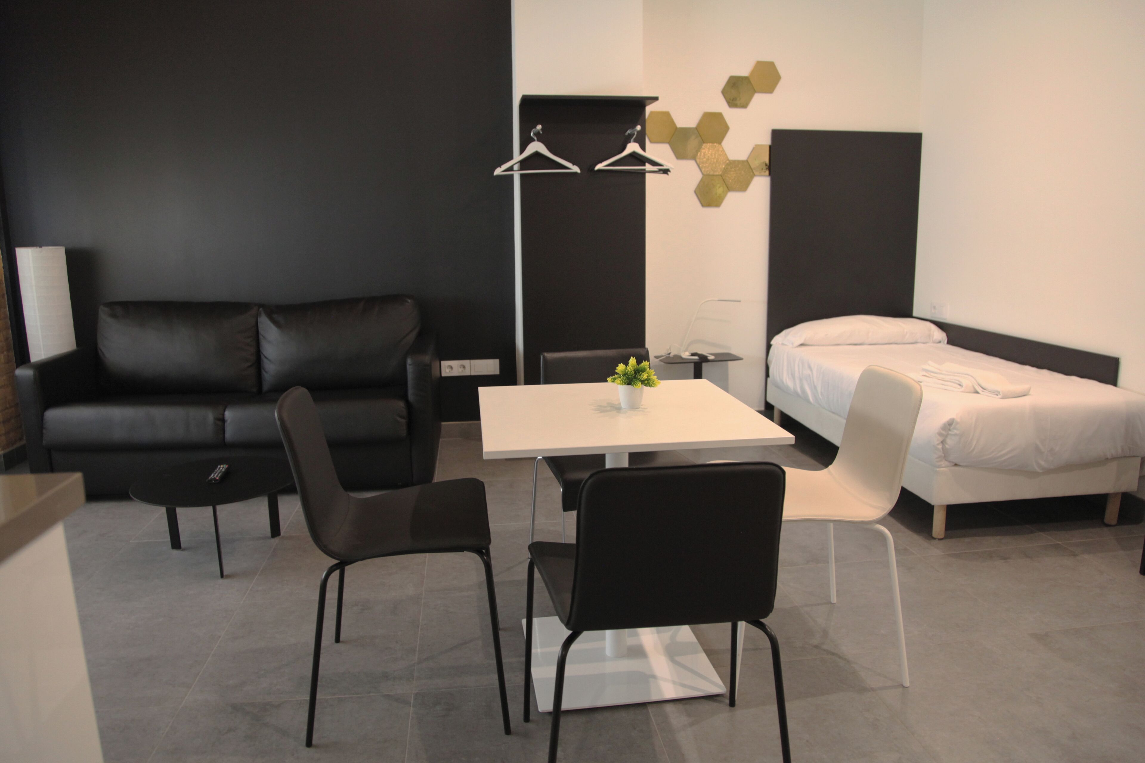 A modern hotel room with a black leather sofa, a white table, two black chairs, a white bed, and a small potted plant.