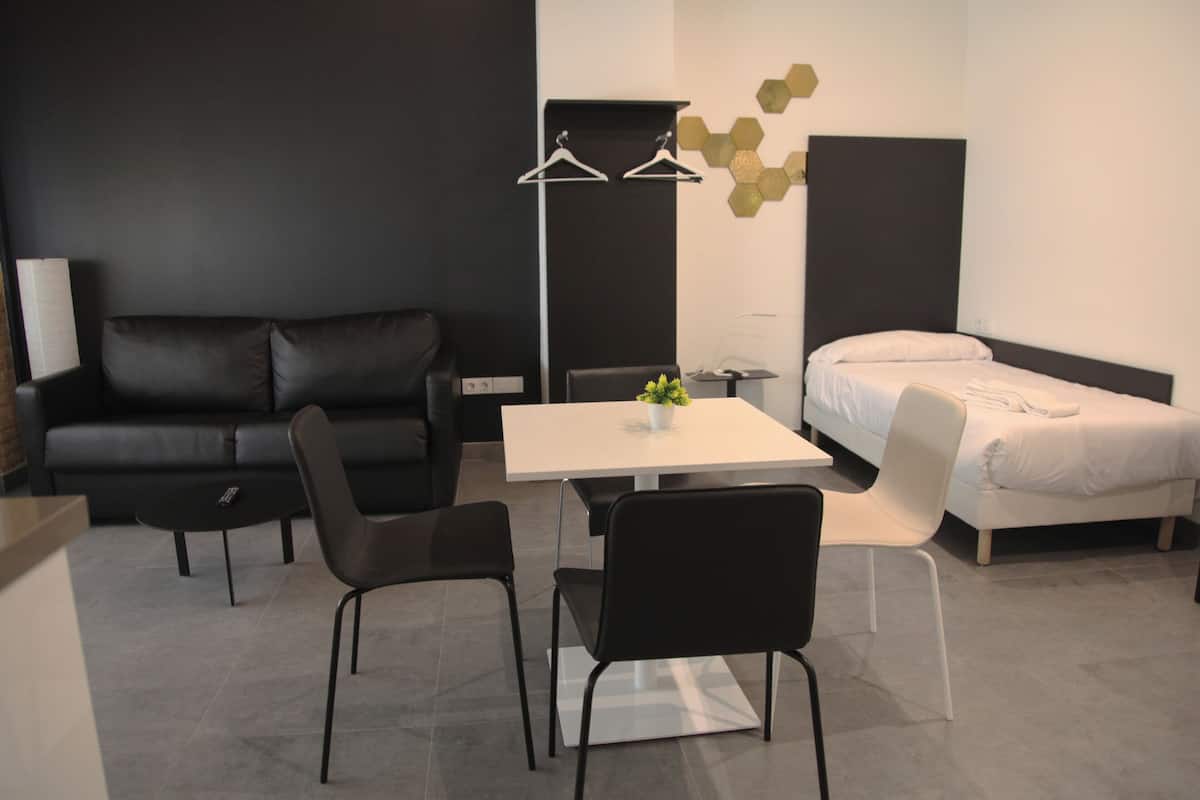 Una habitación de hotel moderna con un sofá de cuero negro, una mesa blanca, dos sillas negras, una cama blanca y una pequeña planta en maceta.