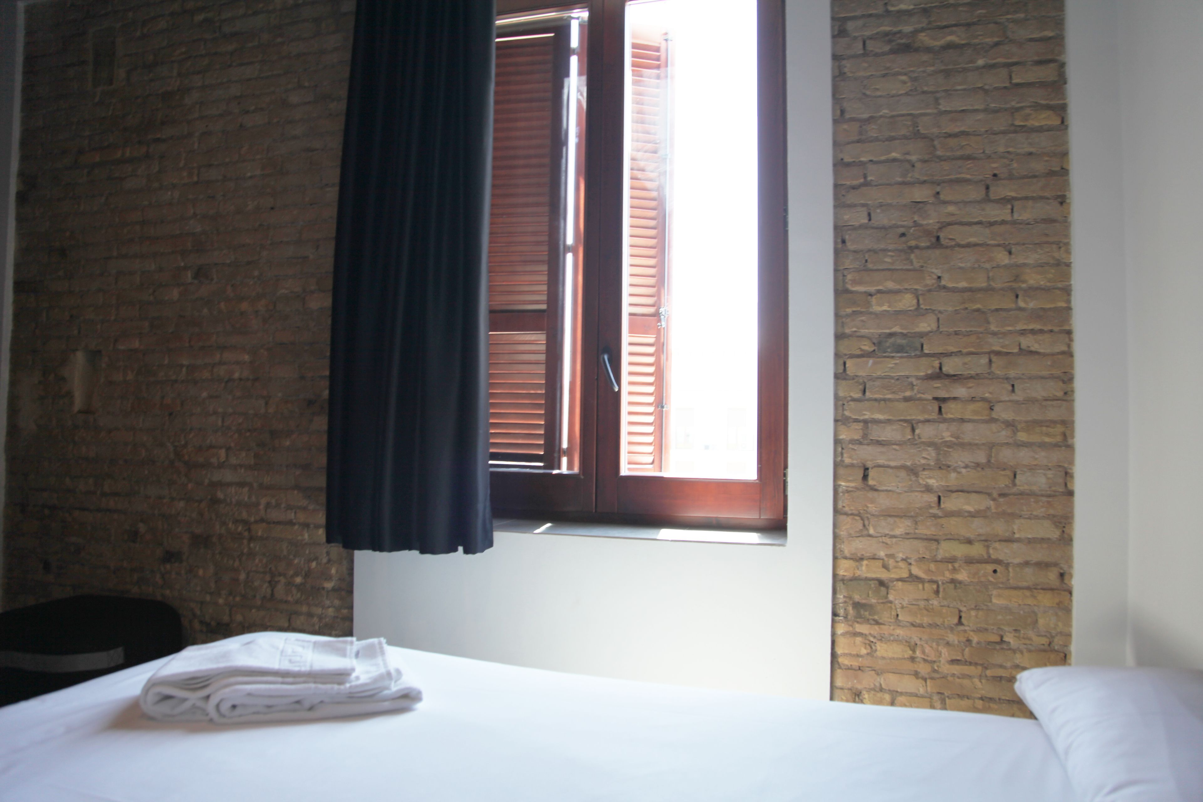 A hotel room with a bed, a window with wooden shutters, and a brick wall.