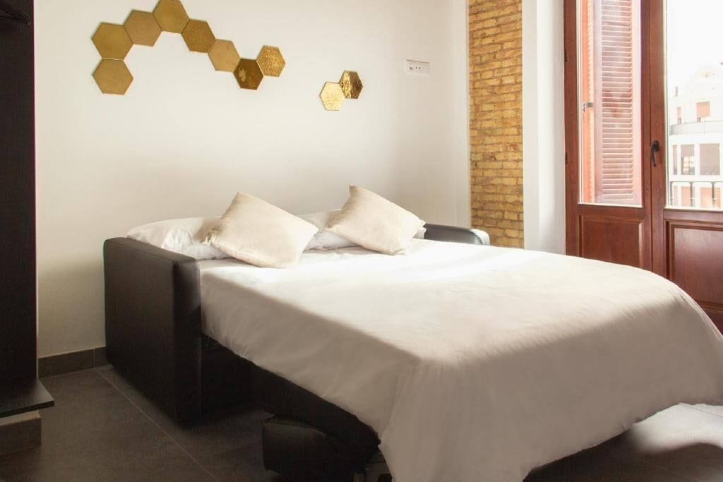 A neatly made bed with white bedding and two pillows, a wall-mounted hexagonal light fixture, and a brick wall.