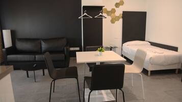 Una habitación de hotel moderna con un sofá de cuero negro, una cama blanca, una mesita con sillas y una mesita de noche con lámpara.