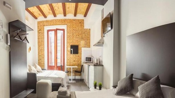 Una habitación de hotel moderna con una cama grande, un sofá, una mesita y una zona de cocina visible a través de una puerta abierta.
