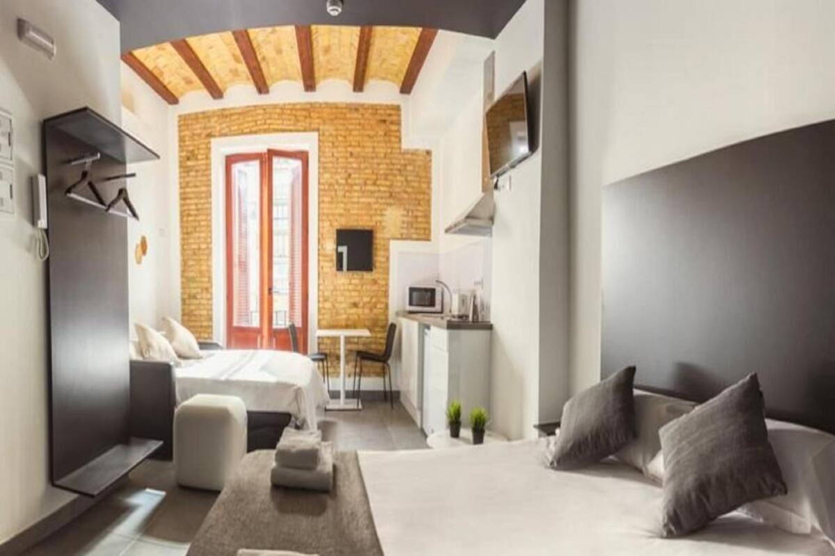 Una habitación de hotel moderna con una cama grande, un sofá, una mesita y una zona de cocina visible a través de una puerta abierta.