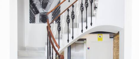 A modern staircase with a unique chandelier design, a white wall with a large abstract painting, and a reception area with a bench.