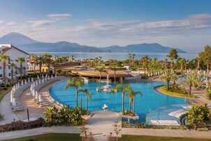 A resort with a large pool, palm trees, and a view of the sea and mountains.