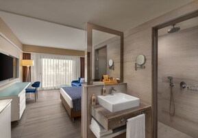 Una habitación de hotel moderna con una cama grande, un escritorio, un baño con lavamanos y espejo, y una ducha.