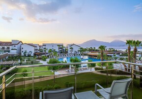 Un balcón de hotel con vista a una piscina, palmeras y una montaña al fondo.