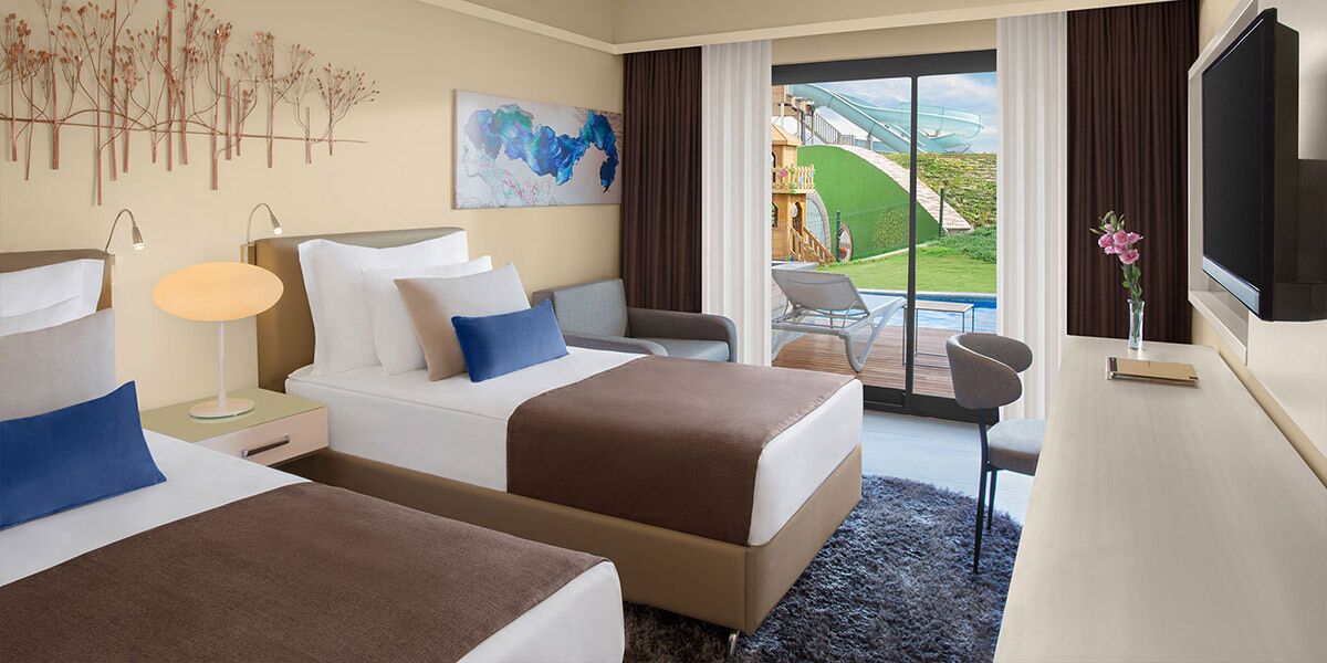 Habitación de hotel con dos camas, televisión, escritorio, silla, lámpara y vista al exterior.