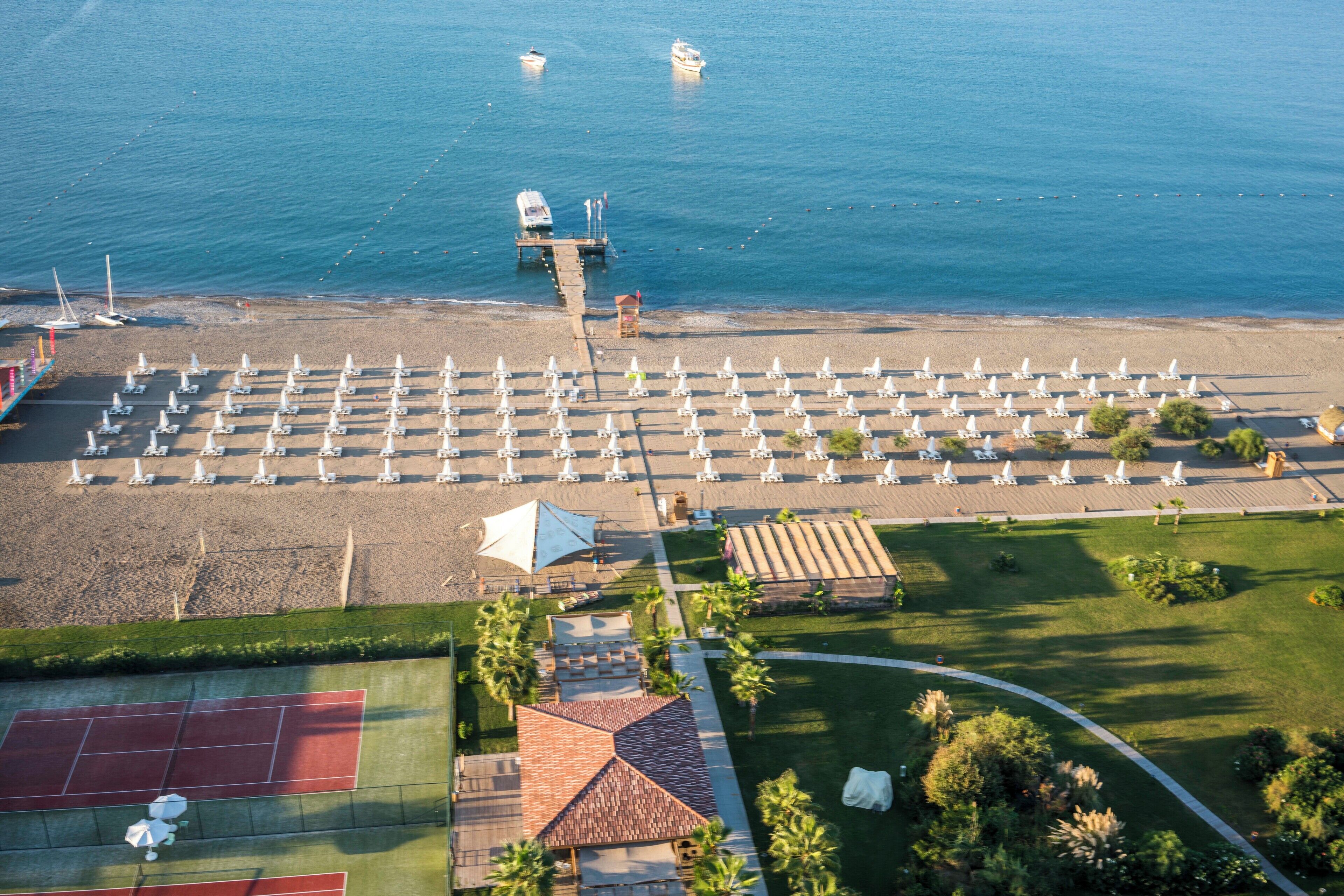 An aerial view of a beach resort with rows of sun loungers, a tennis court, and a pier extending into the sea.