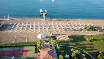 Une vue aérienne d’un complexe hôtelier en bord de mer, avec des rangées de chaises longues, un court de tennis et une jetée qui s’avance dans la mer.