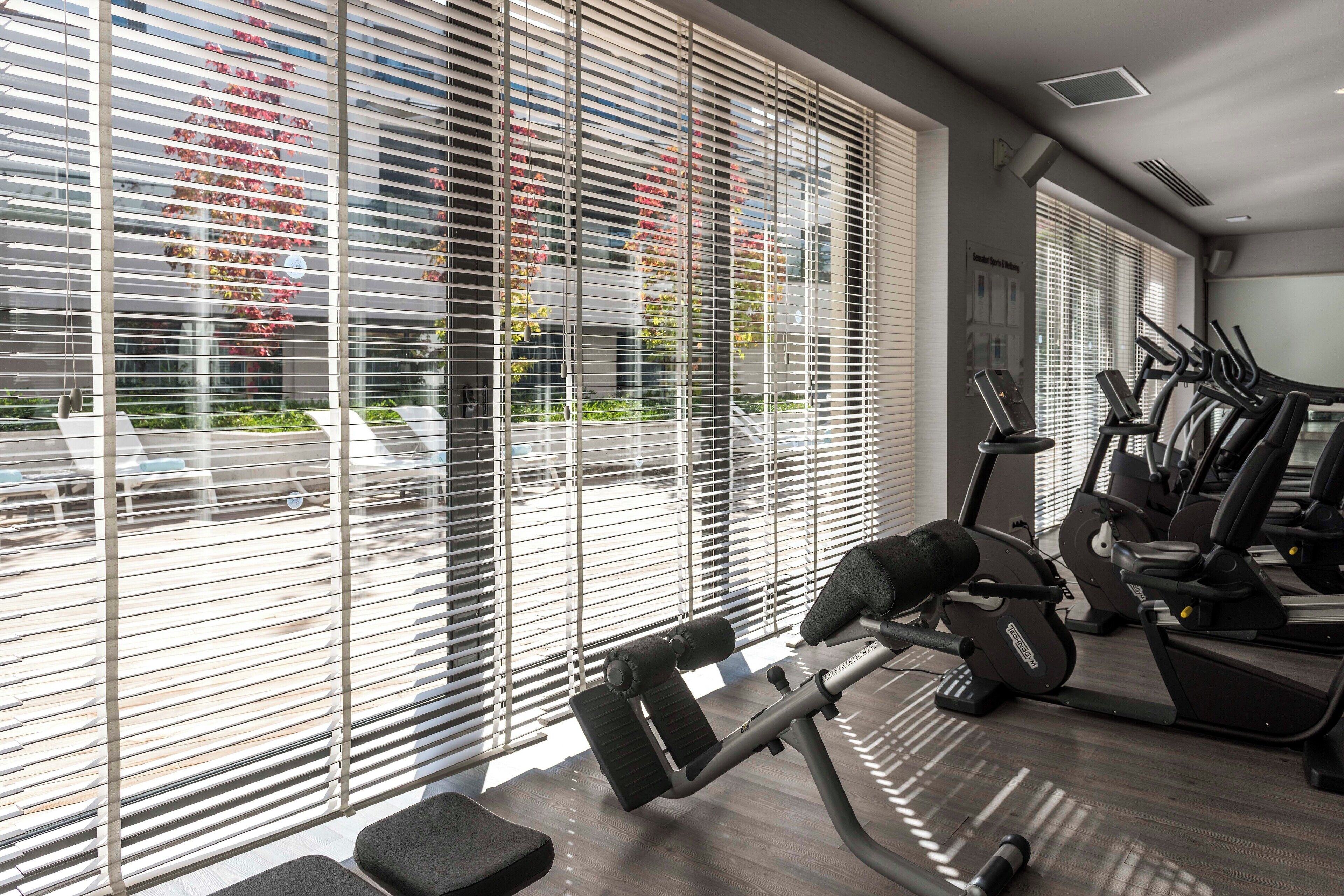 A gym with exercise equipment, large windows with blinds, and a view of the outside.