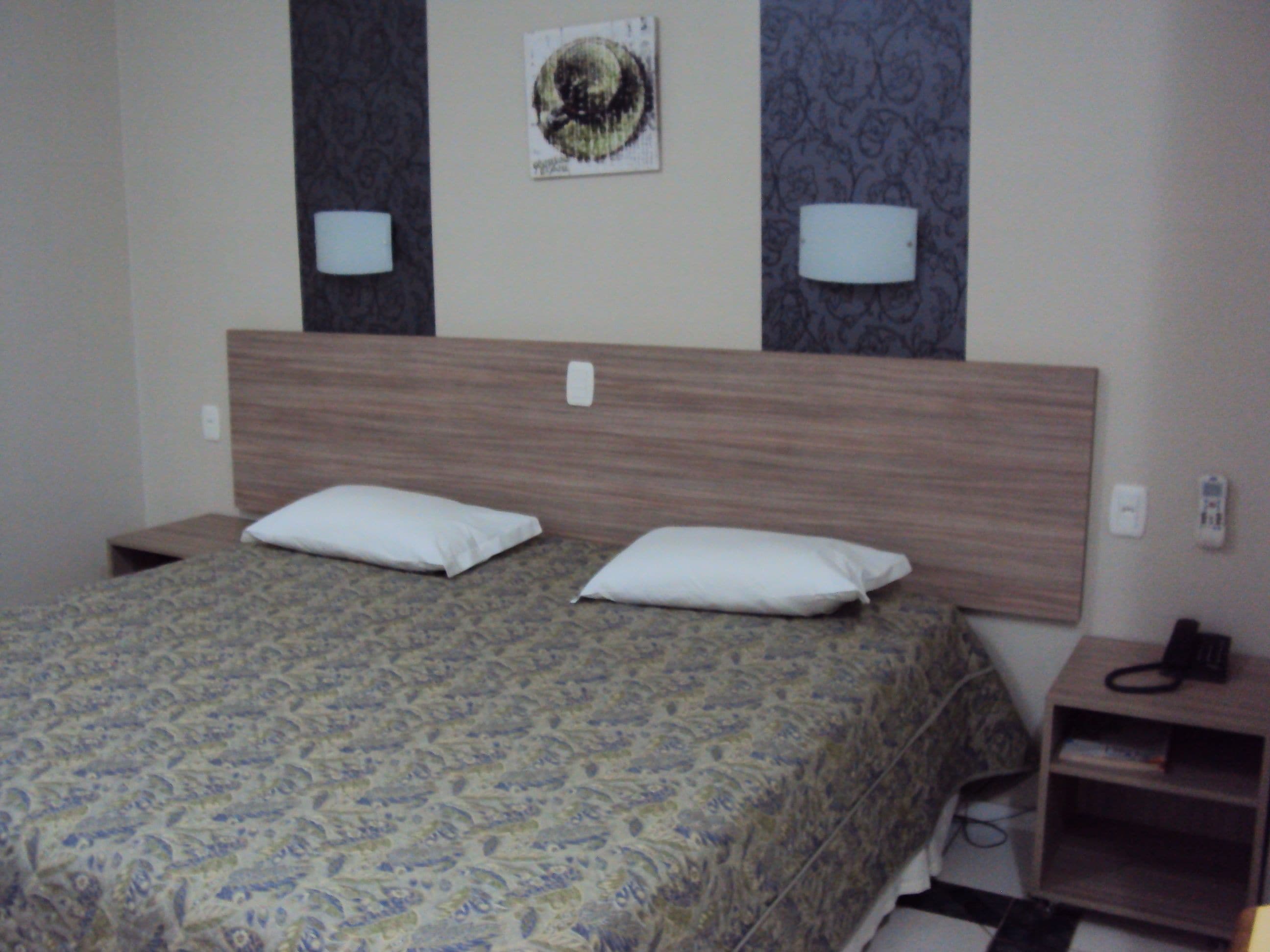 A hotel room with a bed, two pillows, a headboard, two wall-mounted lamps, a bedside table with a phone, and a small shelf.