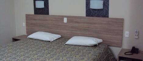 A hotel room with a bed, two pillows, a headboard, two wall-mounted lamps, a bedside table with a phone, and a small shelf.