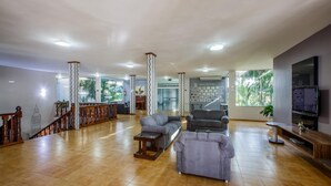 A spacious living area with a high ceiling, a staircase with wooden railings, and a large window offering a view of palm trees. - Olinda Hotel e Eventos (Toledo)