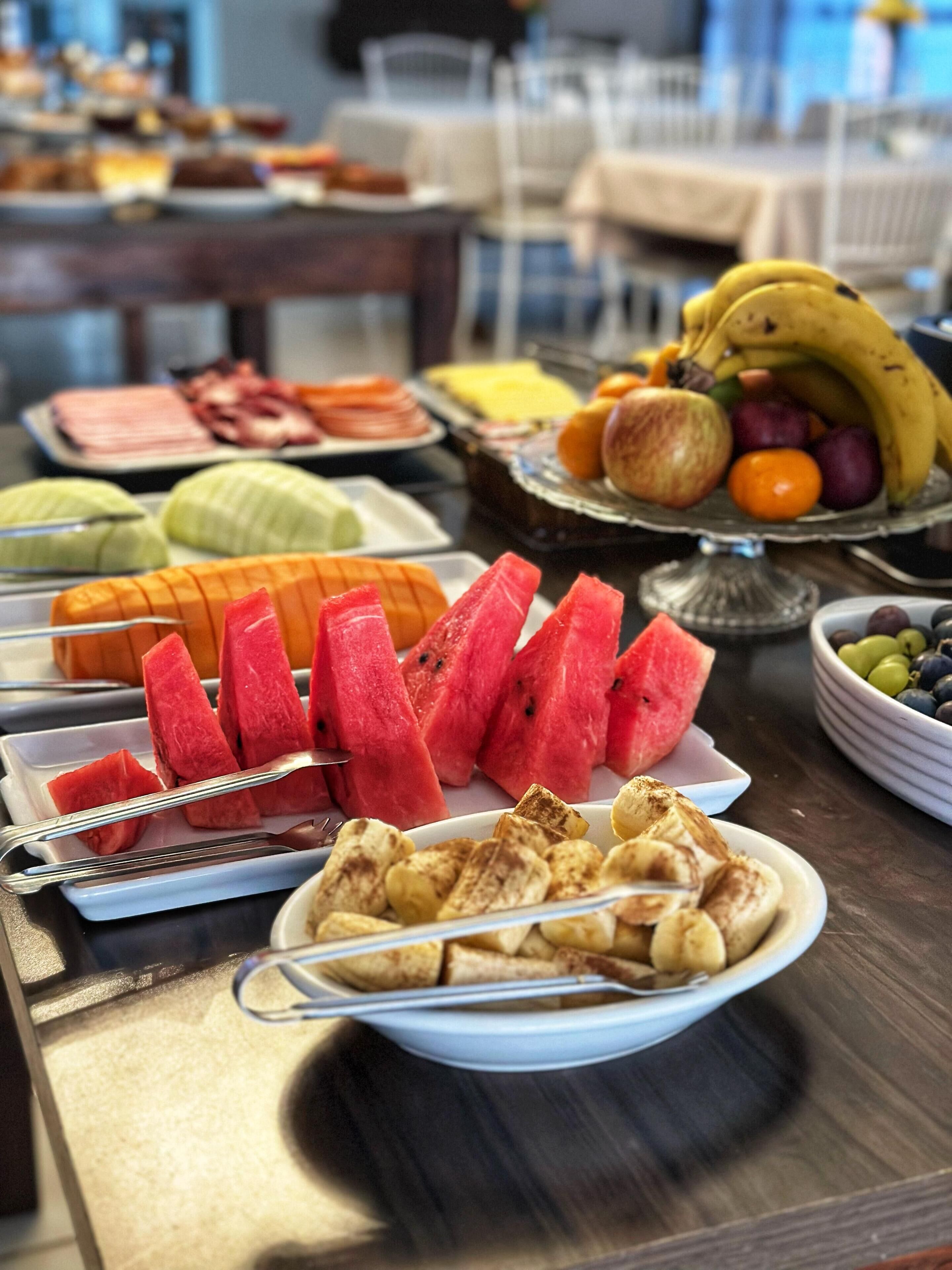 Une table de buffet proposant divers aliments, notamment des pastèques, des bananes et d’autres fruits.