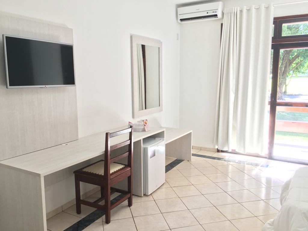 Habitación de hotel con televisión de pantalla plana en la pared, un escritorio con silla, un refrigerador pequeño y una ventana con cortinas.