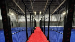 Un pasillo con alfombra roja que conduce a una cancha de tenis azul, delimitada por vallas metálicas.