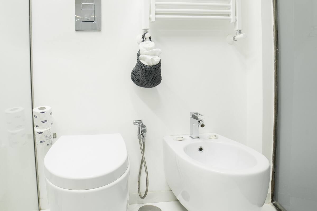 Ein modernes Badezimmer mit Toilette, Bidet und Waschbecken. Regale mit Pflegeprodukten und ein Handtuchhalter sind vorhanden.