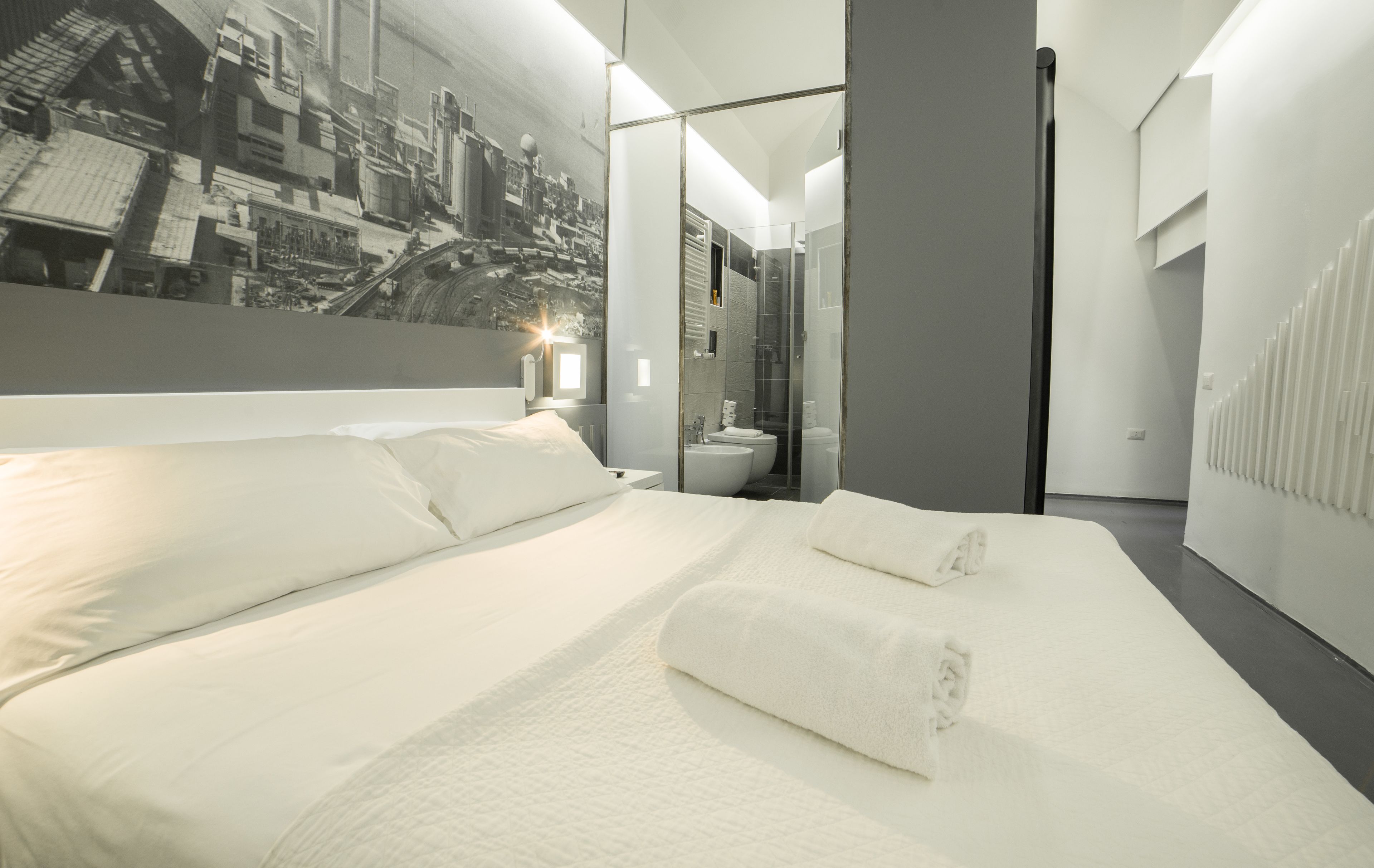 Camera d'albergo moderna con un letto grande, bagno con doccia in vetro e un murale raffigurante una veduta urbana.